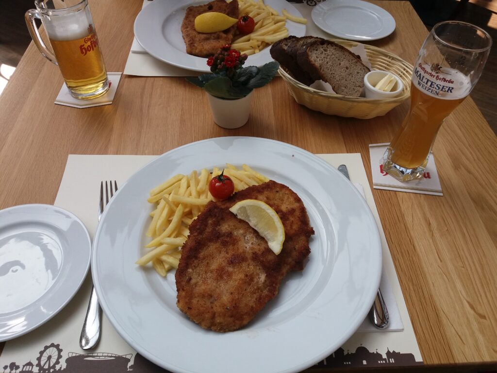 Mesa com um prato servido de schnitzel e batatas fritas, acompanhado de pão e cerveja.